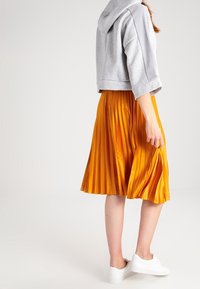 Mujer con un hoodie gris corto, falda plisada naranja brillante y zapatillas blancas de pie frente a un fondo plano.