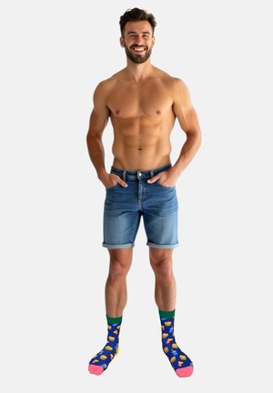 Lächelnder, oberkörperfreier Mann in hochgekrempelten Jeansshorts und bunten Kniestrümpfen mit Essens- und Getränkmustern vor weißem Hintergrund.