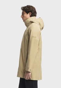Beige wasserdichte Jacke mit Kapuze, die ein elegantes Design und Seitentaschen bietet. Markenlogo auf dem Ärmel sichtbar. Glatte Textur.