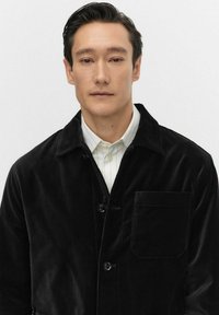 Zwarte corduroy jas met een kraag, knoopsluiting en een borstzak. Gedragen over een wit gestreept overhemd. Gladde textuur.