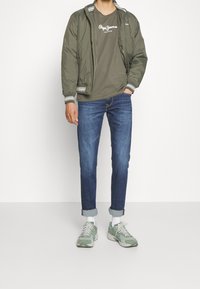 Olivgrön bomberjacka med randiga manschetter, grå grafisk t-shirt, mörkblå jeans och gröna sneakers med vita sulor och accenter.