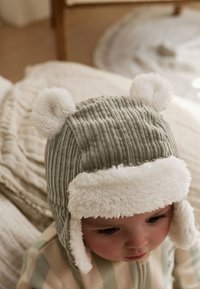 Šedá manšestrová čiapka s bielymi chlpmi na ušiach a mäkkou kožušinovou podšívkou. Zaoblený tvar navrhnutý pre teplo a pohodlie.