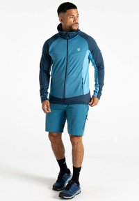 Türkise und marineblaue Sportjacke mit hohem Kragen und Reißverschluss. Kombiniert mit passenden Shorts und schwarzen Socken, die zwei Reißverschlusstaschen haben.