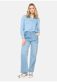 Camicia di jeans blu chiaro con bottoni e tasche sul petto, abbinata a un paio di jeans a gamba larga blu chiaro. Indossata con sneakers bianche e multicolori.