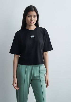 Jeune femme aux longs cheveux foncés portant un T-shirt noir oversize et un pantalon plissé vert sur un fond gris clair.