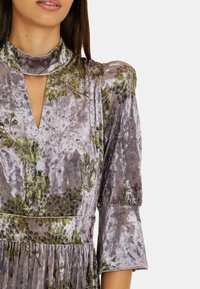 Vestido de terciopelo con un patrón floral en verde sobre una tela gris plateada. Cuello alto con un detalle de ojal y mangas 3/4.