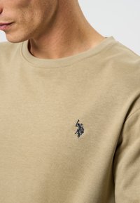 Beige kokvilnas mikina ar apaļu kakla izgriezumu, kurai kreisajā krūtī ir neliels tumši zils izšūts logo. Gluda tekstūra, ikdienišķs dizains.