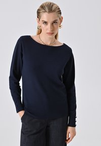 Dunkelblauer Strickpullover mit rundem Ausschnitt, langen Ärmeln und Knopfdetails an den Manschetten. Weiche Textur und taillierte Passform.