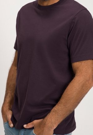 Mann trägt ein einfarbiges dunkelviolettes kurzärmeliges T-Shirt mit Händen in hellblauen Jeans-Taschen, zugeschnitten an Schultern und Hüften.