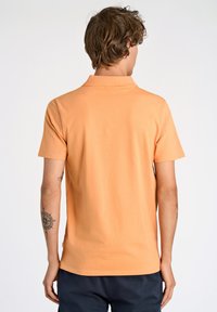 Polo a maniche corte in cotone arancione chiaro con colletto classico e texture liscia, dotato di spacchi laterali e branding sottile.