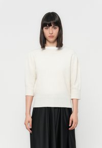 Pull en tricot crème clair avec un col rond et des manches trois-quarts, associé à une jupe fluide noire. Texture douce, coupe décontractée.