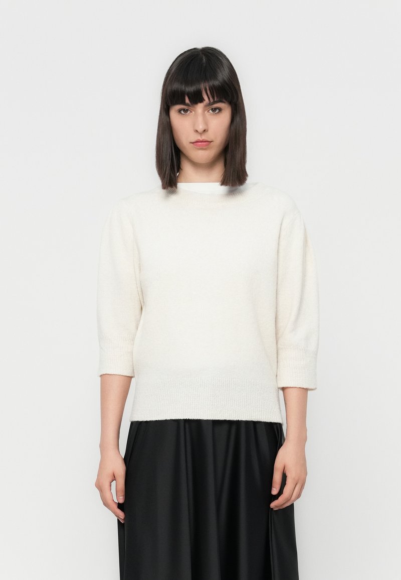 Pull en tricot crème clair avec un col rond et des manches trois-quarts, associé à une jupe fluide noire. Texture douce, coupe décontractée.