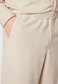Pantaloni iz kordure svetlo bež barve; imajo rebrasto teksturo, elastični pas in stranske žepke. Oblikovanje poudarja udobje in enostavno nošenje.