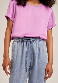 Žena v lesklé růžové blůze s krátkým rukávem, zasunuté do světle modrých jeansů s vysokým pasem a stahovací šňůrkou.