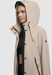 Veste légère beige clair avec capuche, dotée d'une fermeture éclair noire, de boutons-pression et de cordons réglables. Matériau lisse avec un design élégant.