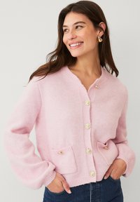 Lichtroze cardigan met een ronde halslijn, vier knopen aan de voorkant en twee voorzakken met gouden accenten. Zachte textuur en een relaxte pasvorm.