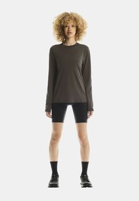 Persoon met krullend blond haar staat rechtop en draagt een donker, langärmige shirt, zwarte fietsshorts, zwarte sokken en zwarte sneakers.