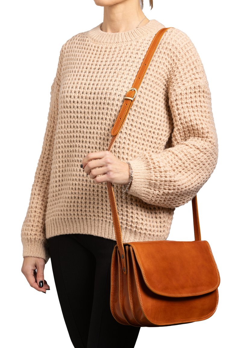 Borsa a tracolla in pelle marrone con pattina piegata, caratterizzata da una texture liscia e una tracolla regolabile. Indossata da una persona con un maglione di lana.