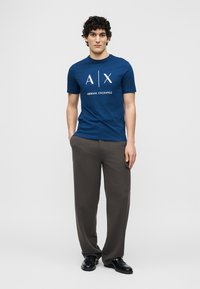 Άνδρας με σγουρά μαλλιά όρθιος, φορά μπλε μπλουζάκι Armani Exchange, σκούρο γκρι παντελόνι και μαύρα παπούτσια, με τα χέρια στις τσέπες.