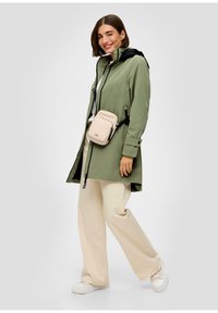 Imperméable léger, vert olive, avec une capuche noire, fermeture éclair et taille ajustable. Associé à un pantalon large crème et des baskets blanches.