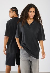 Svart oversized polo med knappelukking på kragen, kombinert med lysegrå baggy jeans. Teksturliknende stoff og avslappet passform. Modellen poserer foran og bak.