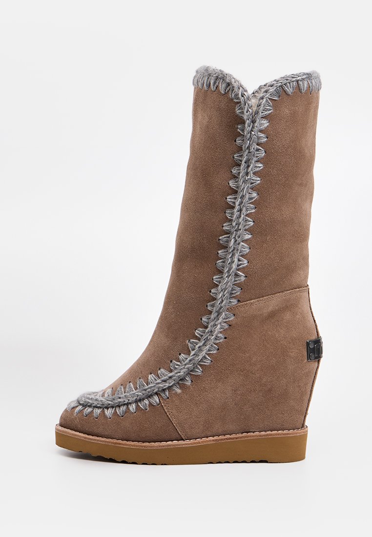 Botas de cuña de ante marrón con costuras grises, un caña alta y una superficie texturizada. Presentan un acento de logo en el lateral y una suela de goma.