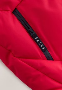 Rote gepolsterte Tasche mit einem schwarzen Reißverschluss und einem Etikett mit der Aufschrift "BAKER." Die Oberfläche ist glatt und hat einen leichten Glanz.