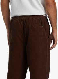 Brown corduroy pants featuring an elastic waistband, side pockets, and a back pocket with a small logo accent. Soft texture with vertical ribs.