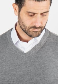 Pull gris à col en V fabriqué en tissu doux, porté par-dessus une chemise blanche à col. Présente un col côtelé et une texture lisse.