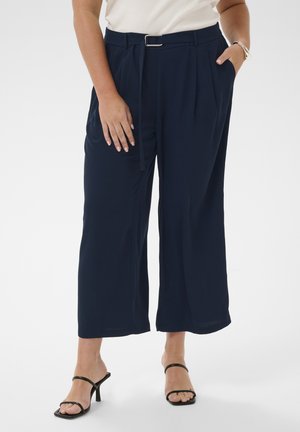 Person trägt marineblaue, hoch taillierte, weite, gekürzte Hose mit Gürtel, weißes Oberteil, silbernes Armband und schwarze Sandalen mit Absatz vor weißem Hintergrund.