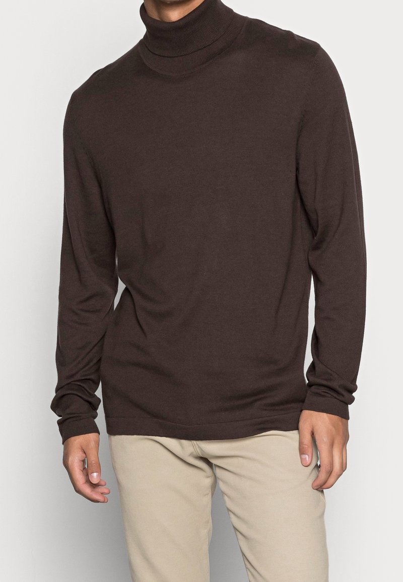Pull en col roulé marron en tissu doux, à manches longues et coupe décontractée, porté par une personne avec un pantalon beige clair.
