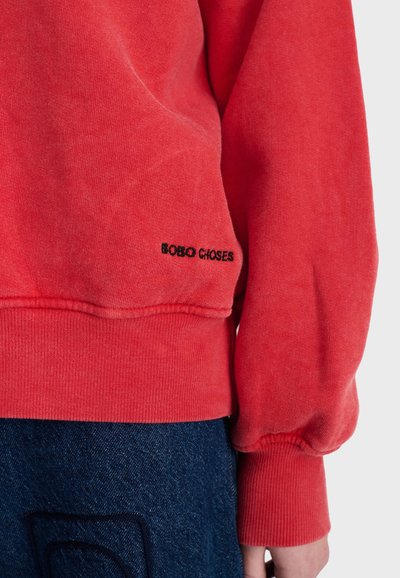 Sweat-shirt rouge à manches longues avec poignets et ourlet côtelés, avec un petit texte noir "BOBO CHOSES" près de la taille, associé à un jean en denim bleu.