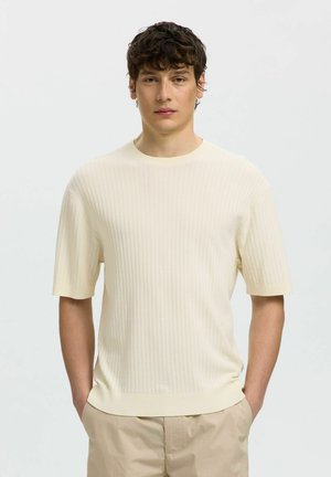 Junger Mann mit lockigem Haar, der einen cremefarbenen, gerippten Kurzpullover und beige Hosen trägt, steht mit den Händen in den Taschen.