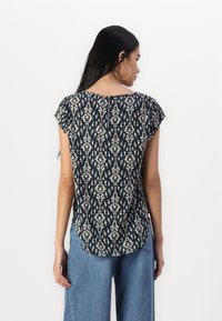 Mørkeblå bluse med kremfarget blomstertrykk, korte ermer, rund utringning og nøkkelhullsdetalj på ryggen. Kombinert med lyseblå bredbeinjeans.