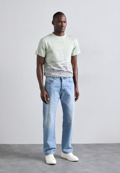 Lys grønn t-skjorte med subtile horisontale stripete gradienter, kombinert med lys blå jeans og hvite joggesko, satt mot en nøytral bakgrunn.