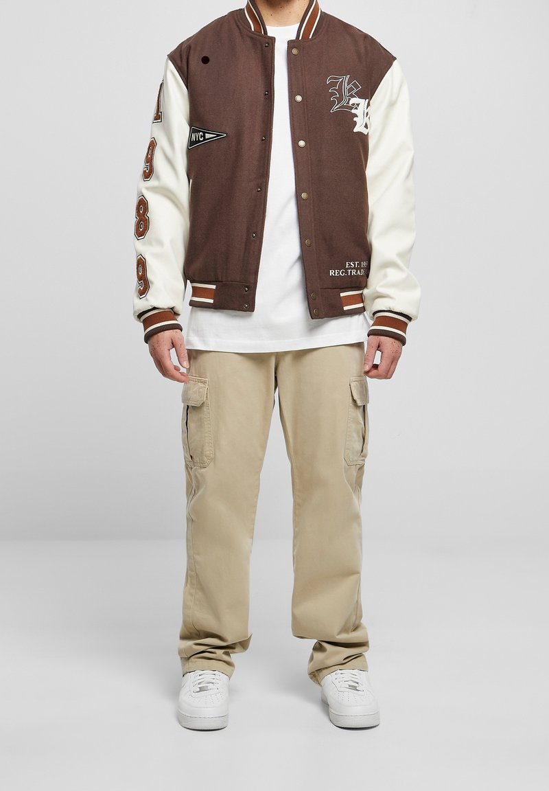 Giacca varsity marrone e crema con toppe ricamate, bottoni a pressione e bordi a righe. Abbinata a pantaloni cargo beige e sneaker bianche.