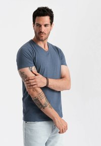 Modrá bavlněná triko s výstřihem do V a krátkými rukávy, které nosí muž; má viditelné tetování na předlohách a uvolněný střih.