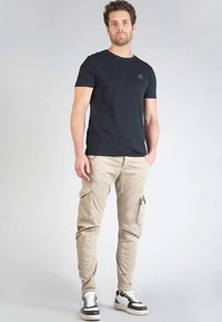 Zwart T-shirt met ronde hals gecombineerd met beige cargobroek met zijzakken. Het model draagt zwart-witte sneakers. Simpel ontwerp, casual pasvorm.