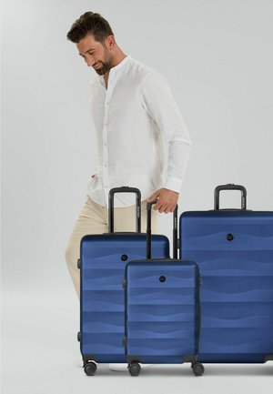 Homme en chemise blanche et pantalon beige debout à côté de trois valises bleues avec poignées extensibles sur un fond uni.