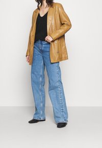 Brun läderöversized blazer, svart V-ringad topp, högmidjade ljusblå jeans och svarta ankelstövlar; synliga knappar på blazern.