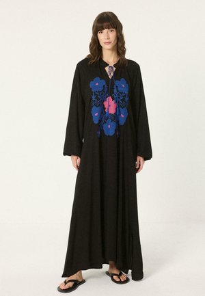 Nő hosszú fekete ruhában, kék és rózsaszín virágos hímzéssel, hosszú ujjú, fekete szandállal, sima háttér előtt áll.