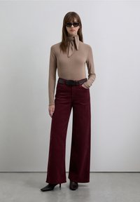 Bluză maro cu mâneci lungi, prietenă, cu un fular legat la gât, asortată cu pantaloni largi bordo și o curea neagră. Ochelarii de soare negri completează look-ul.