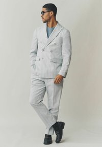 Hellgrauer Nadelstreifenanzug mit einer doppelt geknöpften Jacke und Bundfaltenhosen, kombiniert mit einem blauen Hemd und schwarzen Slippern.