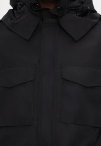 Gros plan d'une veste noire avec deux poches à rabat sur le devant et un col montant avec des détails de boutons.