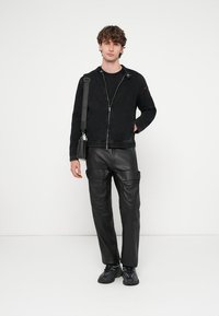 Melna džinsa jaka ar rāvējslēdzēja aizdari, melnas ādas bikses un melni sporta apavi. Jakā ir stāvošs apkaklis un sarkani akcenti.