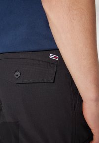 Närbild av en persons höft som bär svarta shorts med en knäppt bakficka och en liten rektangulär röd, vit och blå logotyp.