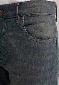 Farkkuhousut haalistuneessa sinisessä värissä, suoran lahkeen muotoilulla, jossa on etutasku, kuparinvistiset niitit ja ruskeita ompeluaksentteja.