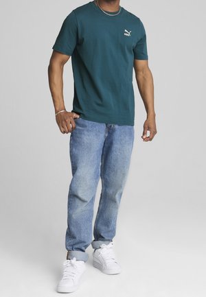 Türkisfarbenes Baumwoll-T-Shirt mit kurzen Ärmeln und kleinem weißem Puma-Logo, kombiniert mit hellblauen Denim-Jeans und weißen Turnschuhen.