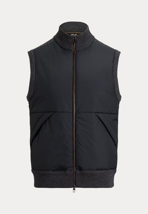 Mann mit runden Sonnenbrillen, dunkle Reißverschlussweste über dunkelgrauem Pullover, schwarze Hose, weiße Schuhe, stehend vor einem einfarbigen beigen Hintergrund.