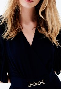 Vestido envolvente azul marino con mangas cortas plisadas, que presenta un detalle de cinturón con acento dorado. Textura de tela suave y diseño con escote en V.